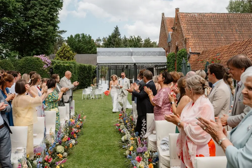 Gasten applaudisseren voor het bruidspaar tijdens een ceremonie in de tuin, met de orangerie en de kasteelhoeve op de achtergrond