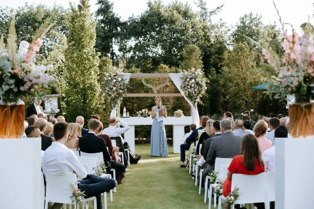 Huwelijksceremonie in de tuin met witte stoelen, bloemstukken en een spreker onder een bloemenboog