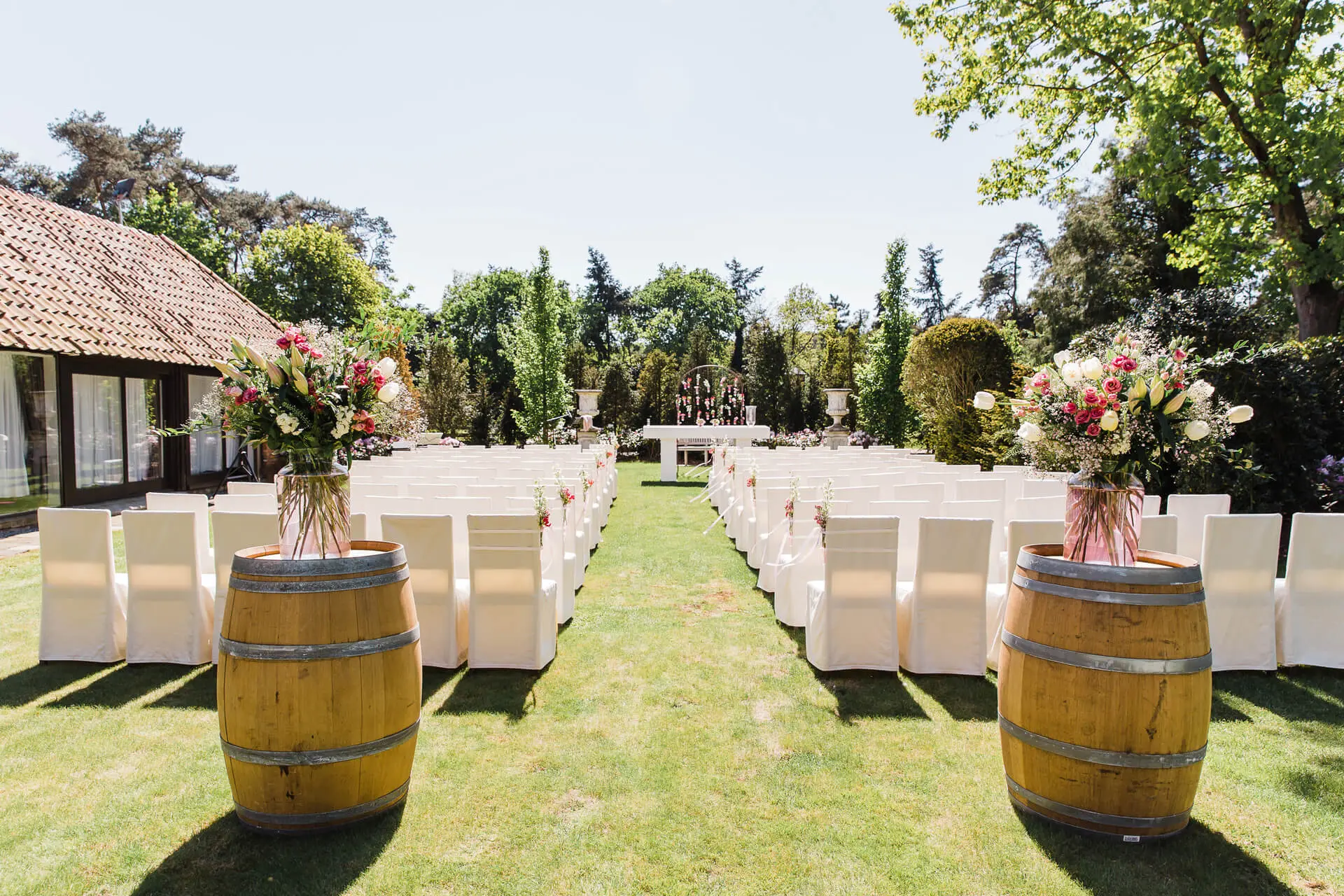 Tuin opgesteld voor een ceremonie met witte stoelen en boeketten op wijntonnen