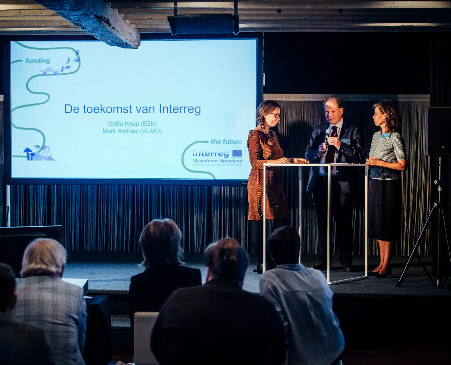 Drie sprekers aan een hoge tafel naast het presentatiescherm