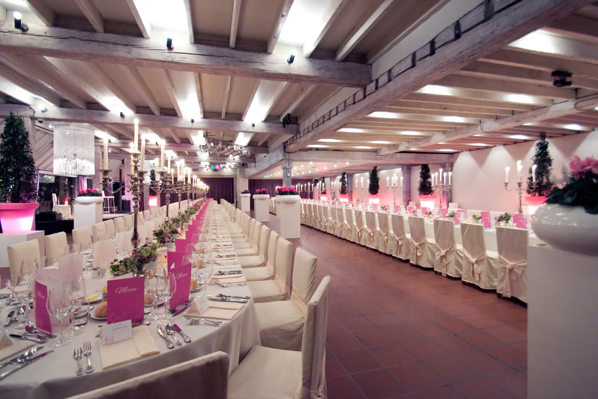 Lange rijen gedekte tafels met witte stoelen en roze accenten in de grote zaal