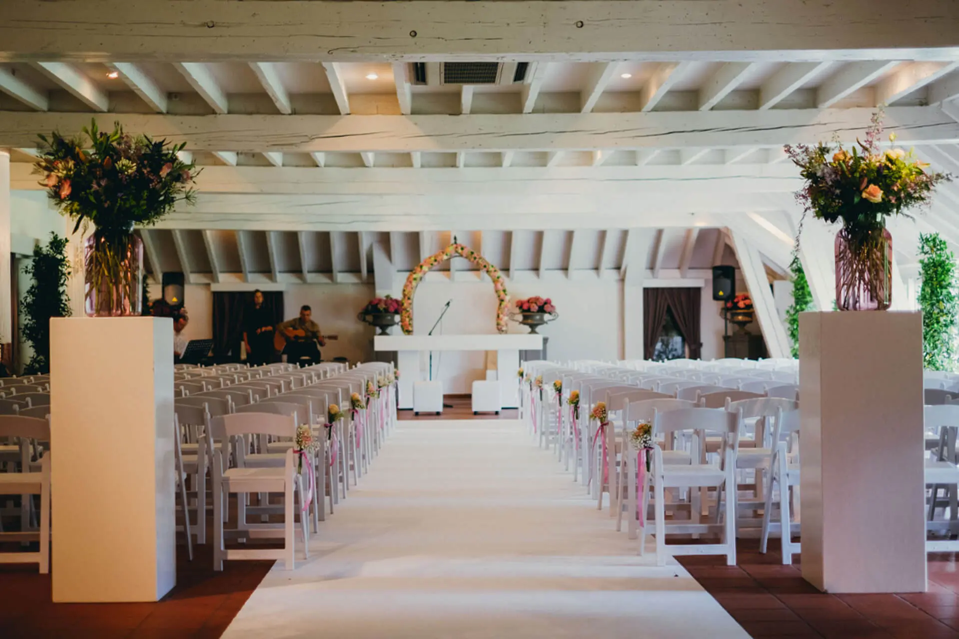 Kleine zaal opgesteld voor een ceremonie met witte stoelen, een loper en hoge boeketten op zuilen