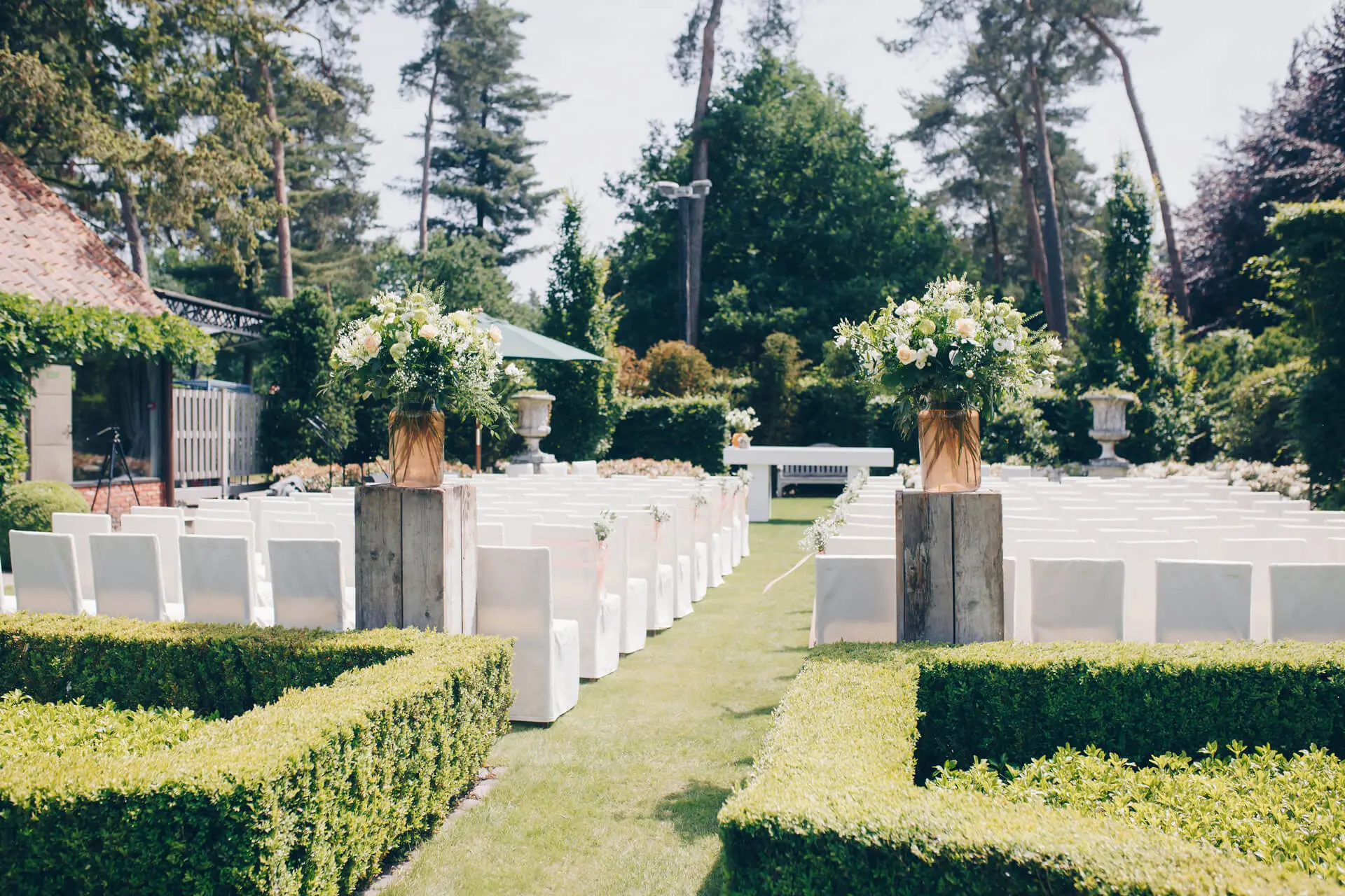 Tuin opgesteld voor een ceremonie met witte banken tussen buxushagen en grote boeketten