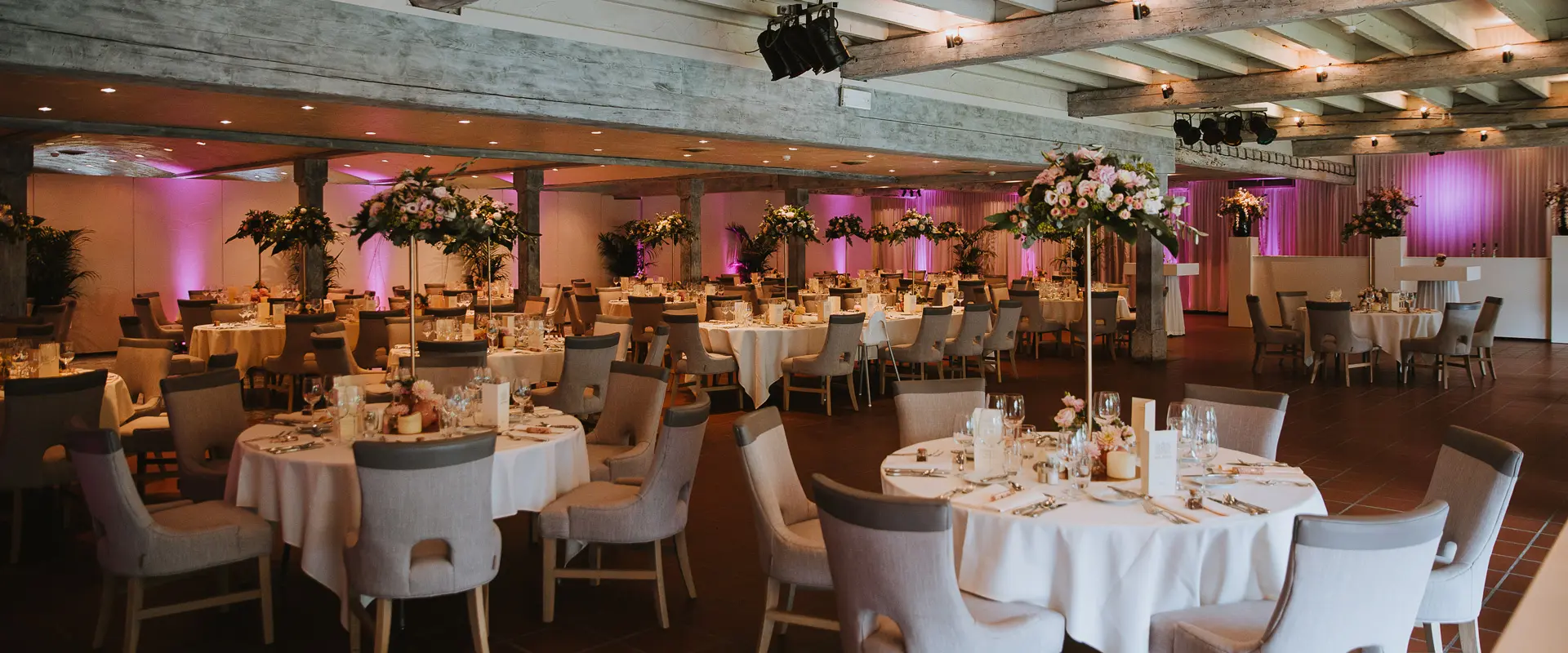 Grote zaal gedekt voor een huwelijksdiner met ronde tafels, witte stoelen en roze sfeerverlichting