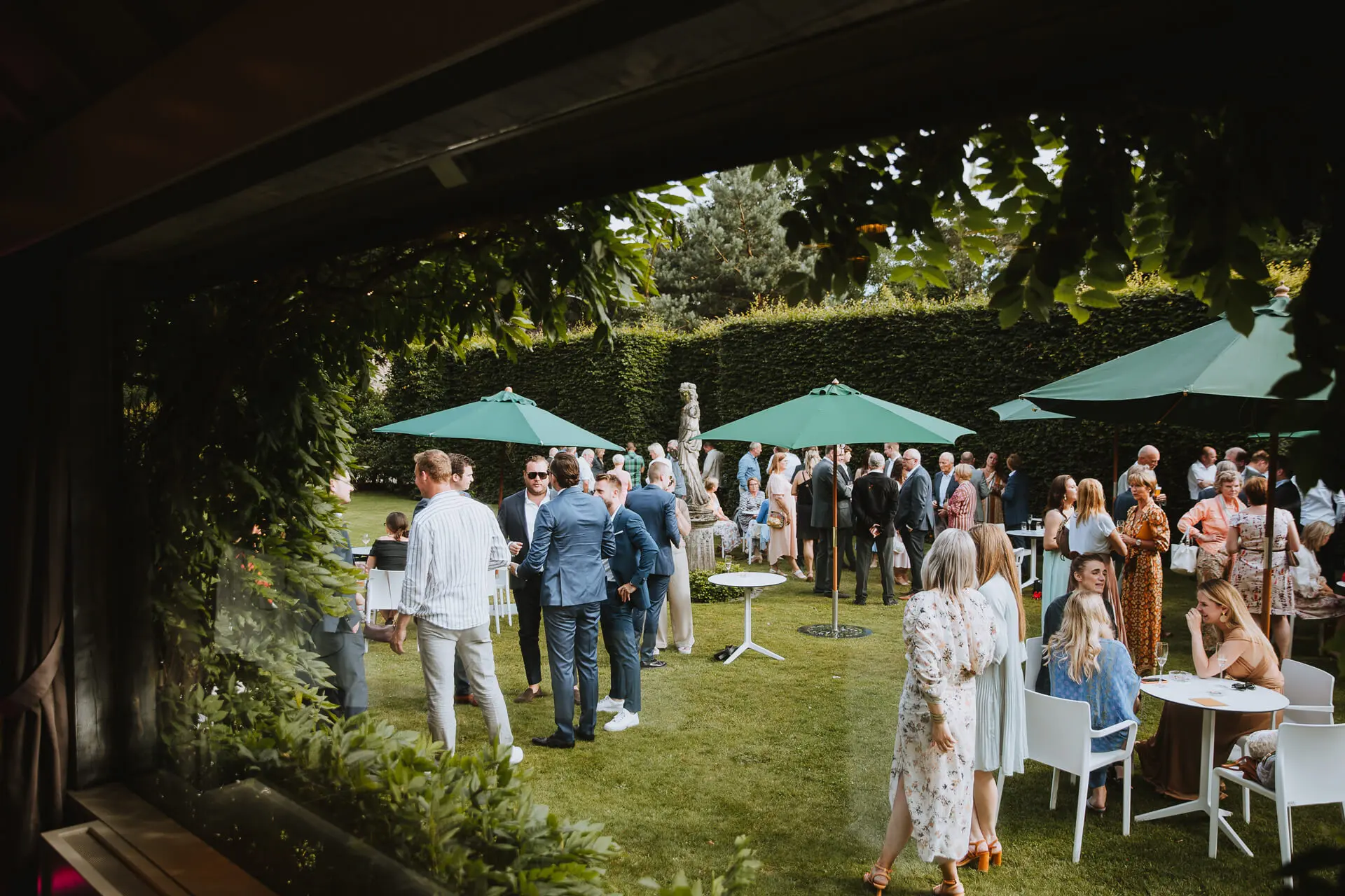 Uitzicht vanuit de zaal op gasten in de groene tuin met parasols
