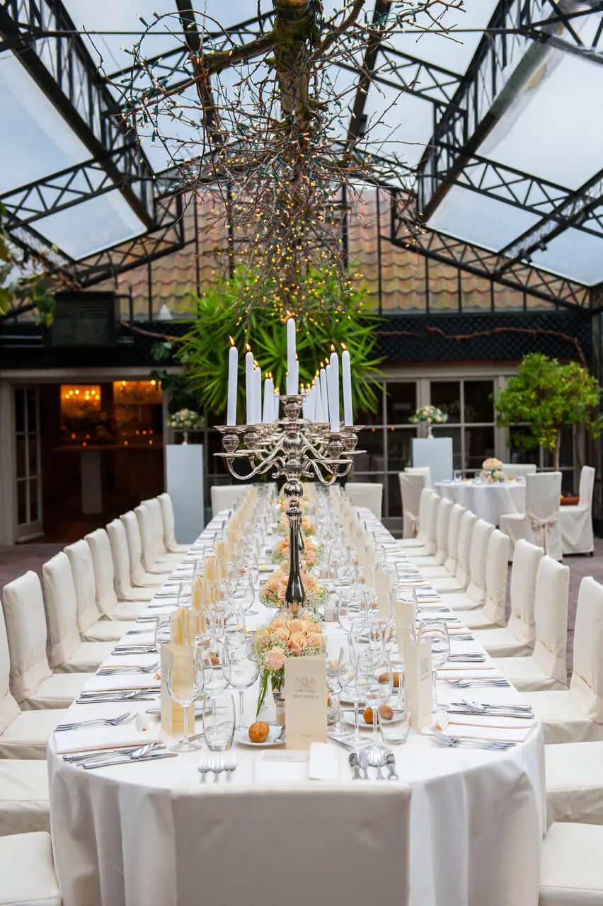 Lange feesttafel met kandelaars en witte stoelen in de orangerie van de grote zaal