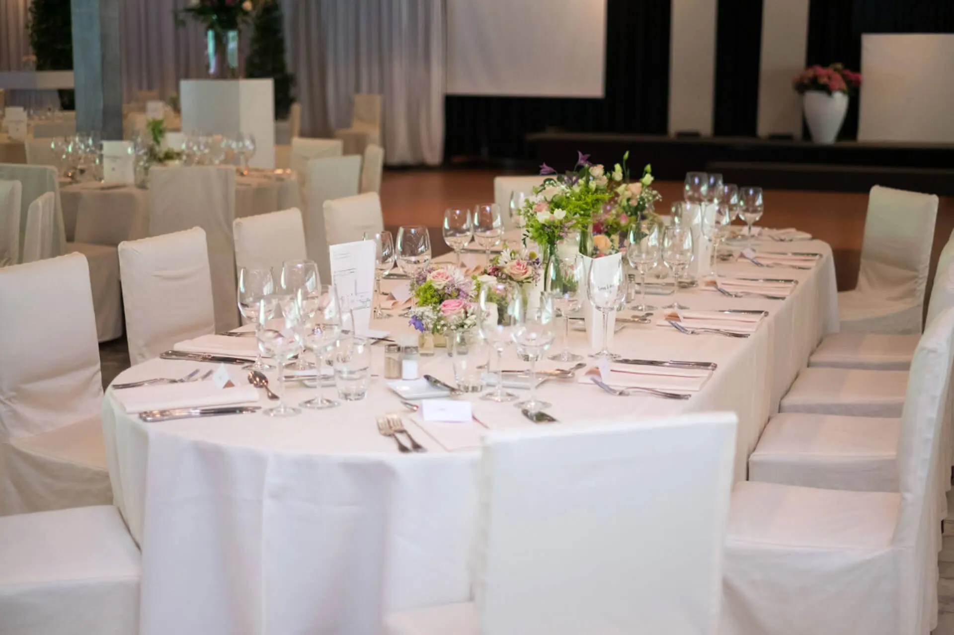Ovale eretafel met wit linnen, glazen en bloemstukjes, omringd door witte stoelen