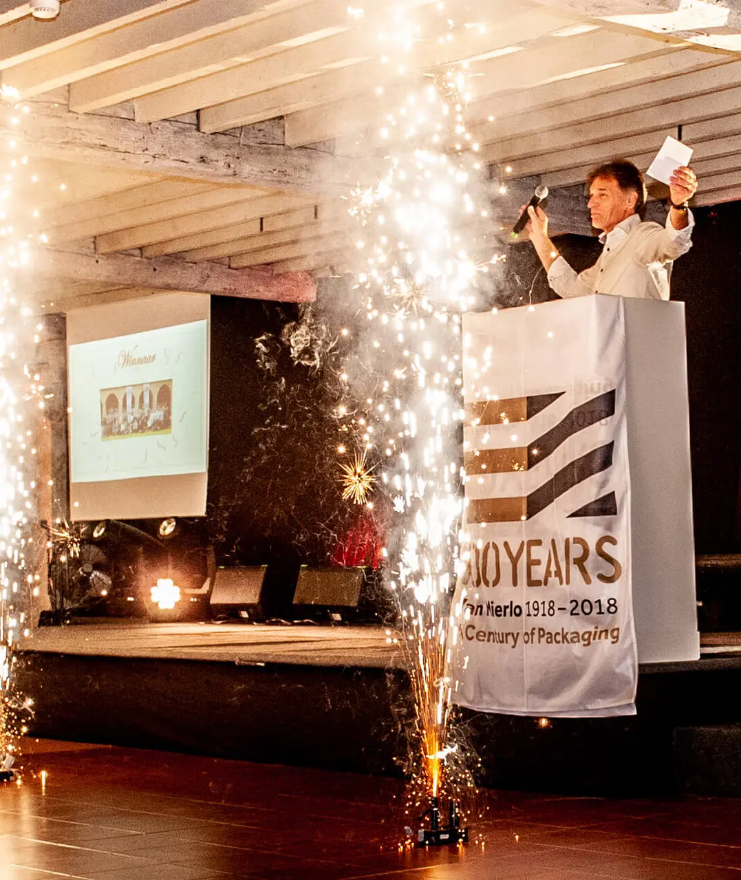 Spreker op het podium tussen vuurwerkfonteinen tijdens een bedrijfsevent bij Alta Ripa