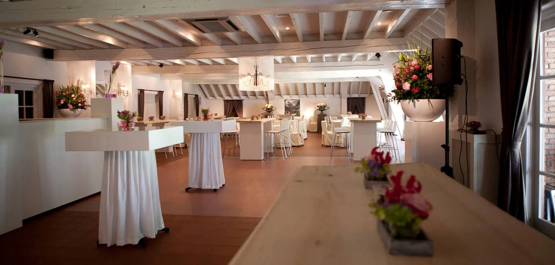 Kleine zaal met hoge tafels in wit linnen en een lange houten tafel met bloemen