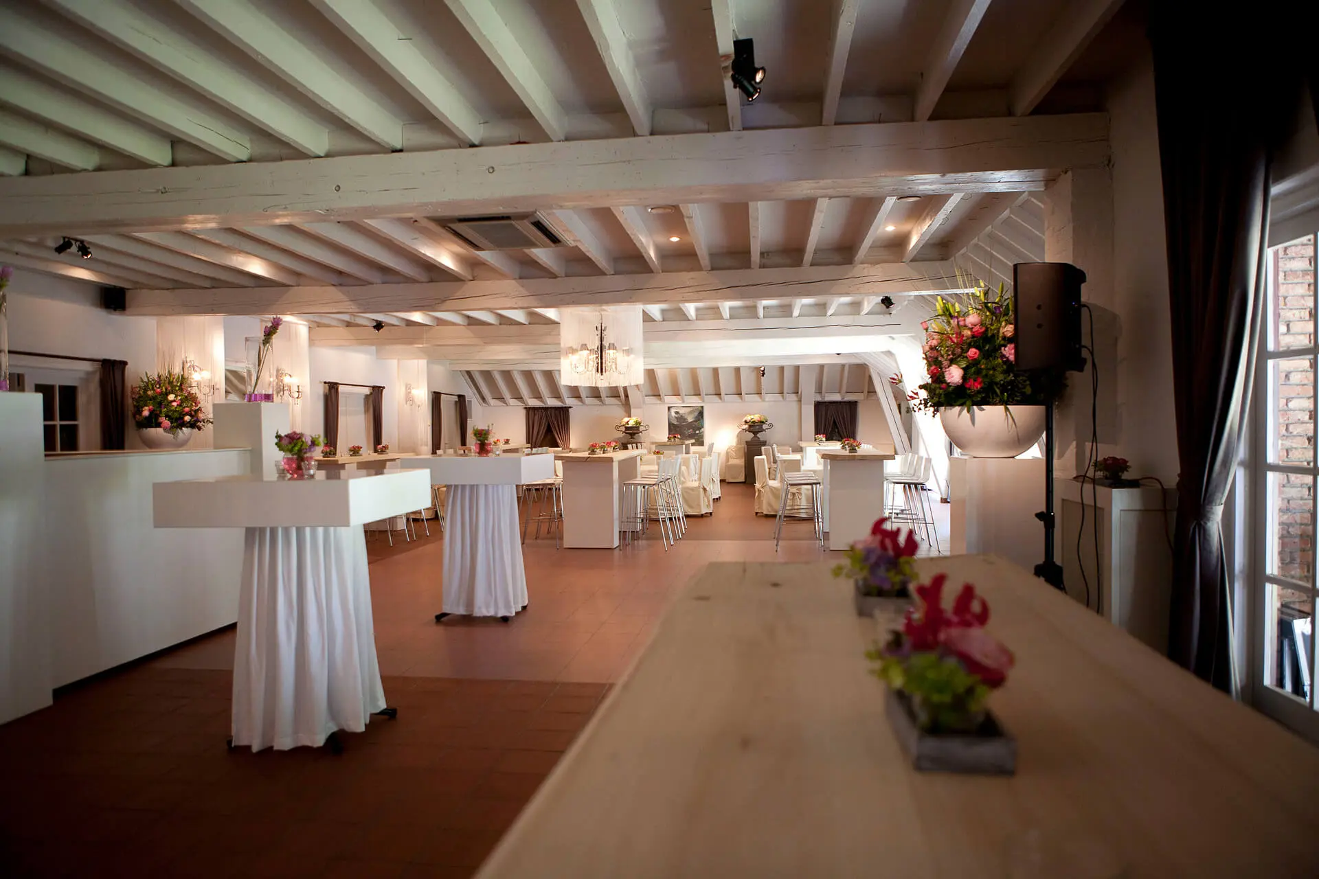 Kleine zaal met hoge tafels in wit linnen en een lange houten tafel met bloemen