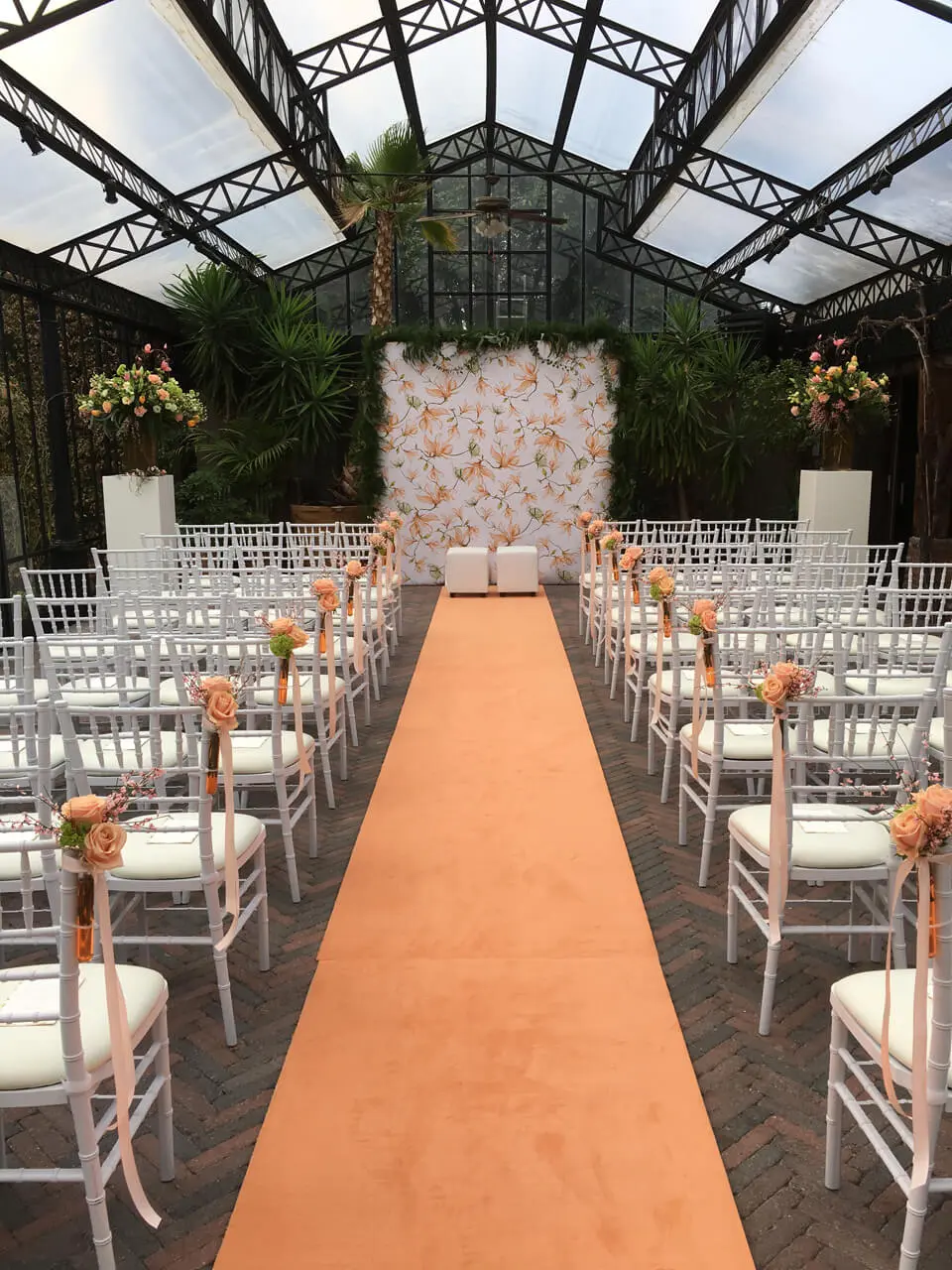 Orangerie opgesteld voor een ceremonie met witte stoelen, een zalmkleurige loper en een bloemenwand