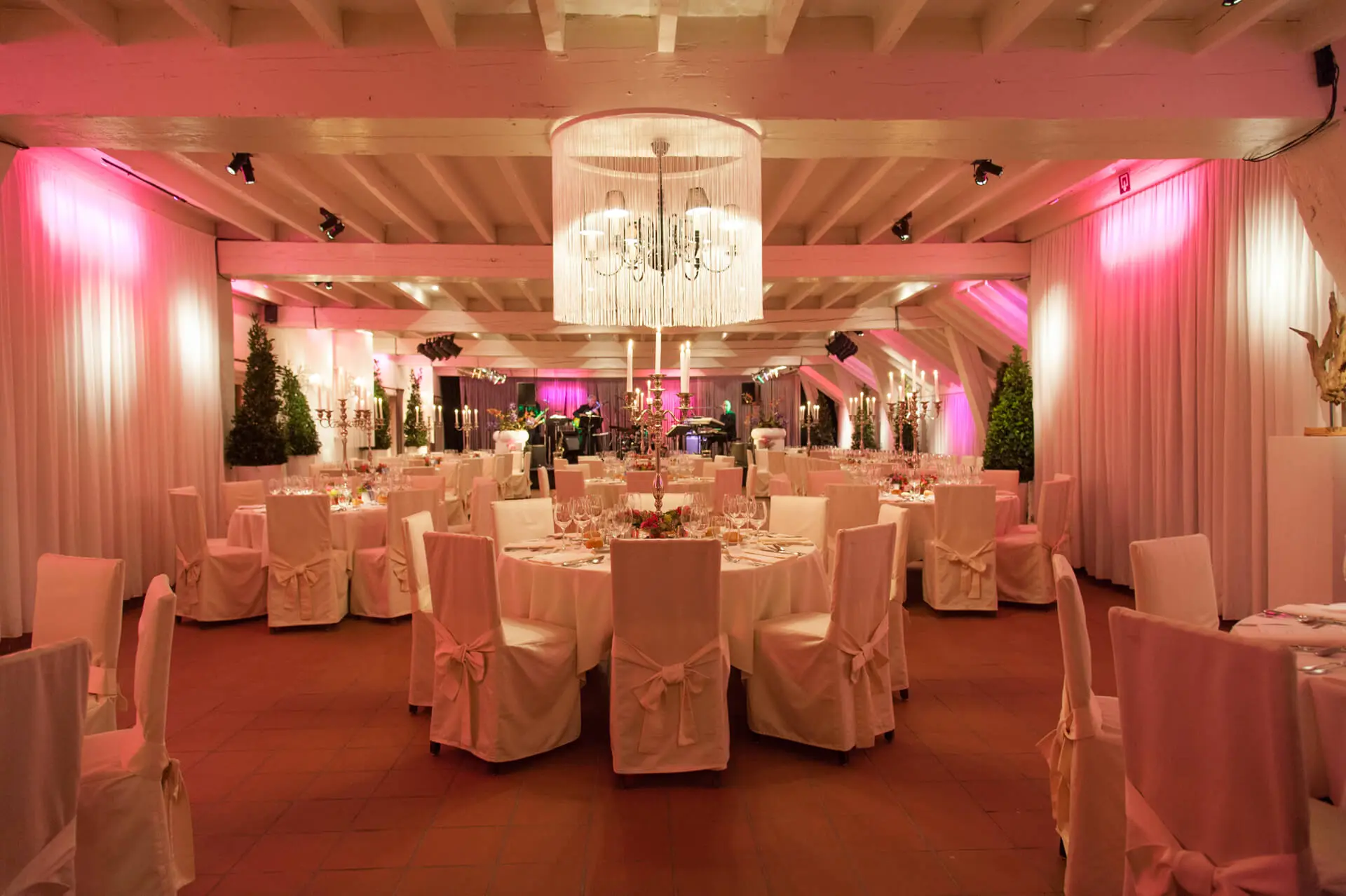 Kleine zaal met ronde tafels, stoelen in witte hoezen en roze sfeerverlichting