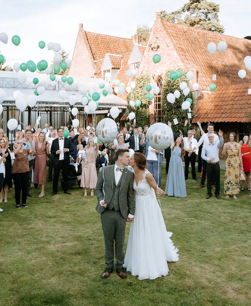 Bruidspaar kust in de tuin terwijl de gasten witte en groene ballonnen oplaten voor de kasteelhoeve